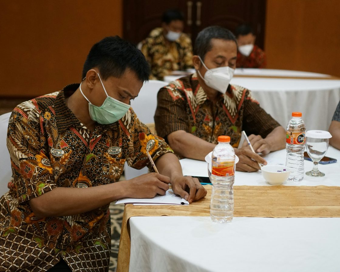 Dua pria berbatik menulis di meja rapat, membandingkan bentuk badan usaha