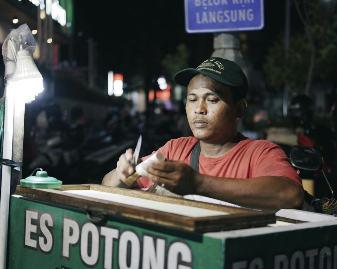 Pedagang es potong menyiapkan dagangan di gerobaknya pada malam hari, gambaran usaha kecil yang memakai tarif final