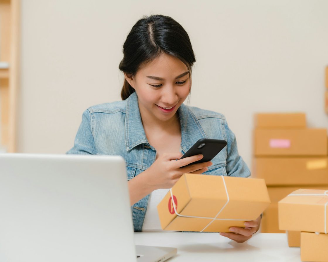 Penjual daring memeriksa paket sambil memegang ponsel di depan laptop